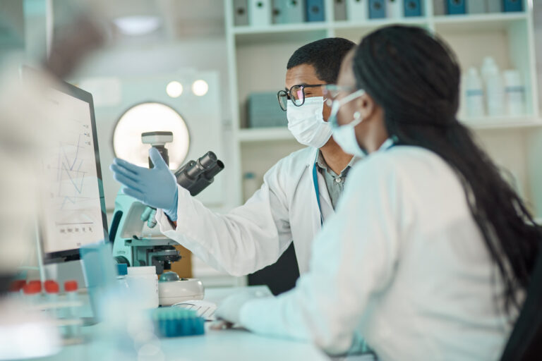 two people seated at a lab desk featuring a pc screen and microscope