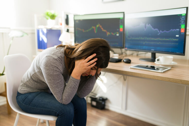 person seated at a desk with two pc monitors holding head in hands