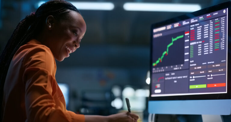 a smiling person writing notes while looking at stock charts on the computer