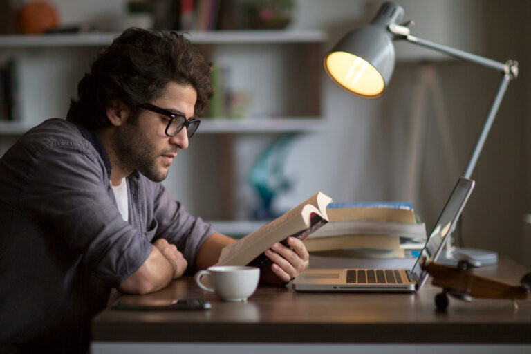 a person sitting at a table with a laptop and a lamp reading a book